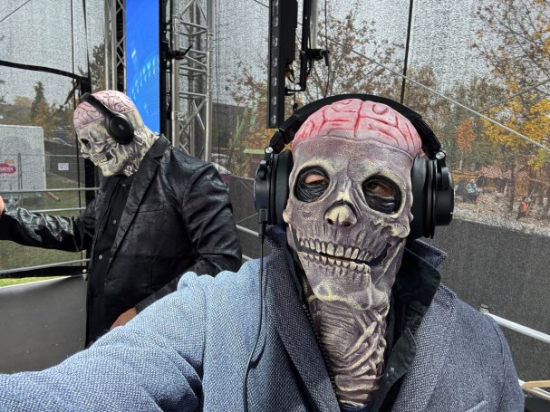 Zwei Personen mit Schädelmasken und Kopfhörern, einer in einem Anzug, stehen im Hintergrund.