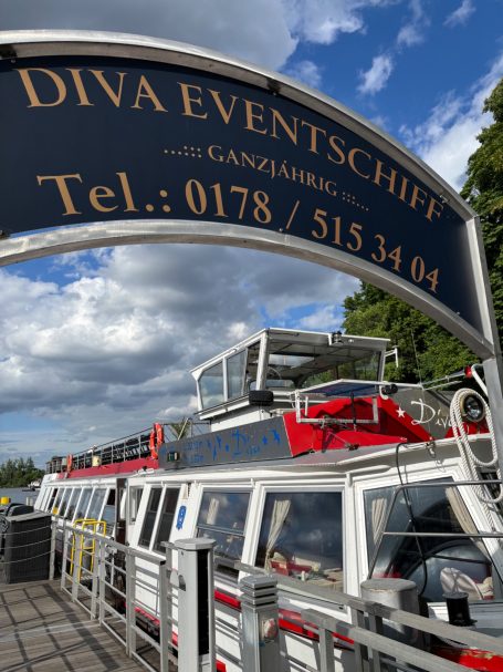 Eventschiff MS Diva Ein Schiff mit dem Namen "Diva" im Hafen, unter einem Schild mit Kontaktdaten.