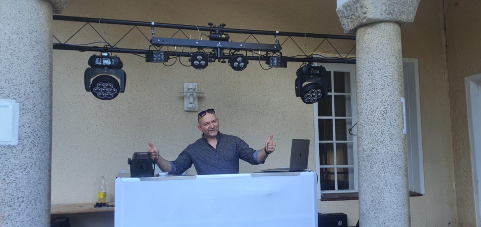 DJ steht hinter einem Mischpult, mit Lichtern im Hintergrund, lächelt und hebt die Arme.