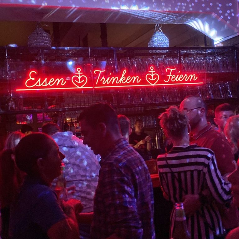 Neon-Schild mit den Worten „Essen, Trinken, Feiern“ in einer Bar mit Menschen.