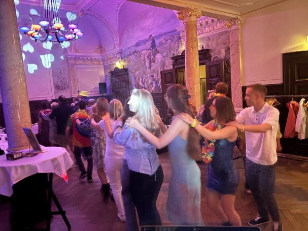 Personen tanzen in einer festlichen Umgebung mit bunten Lichtern und Regalen im Hintergrund.