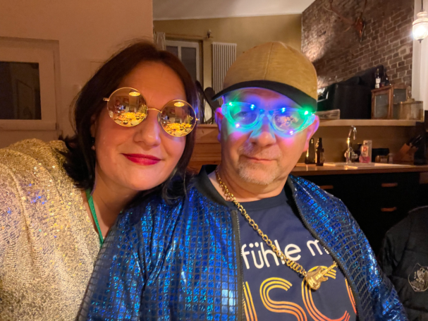 Zwei Personen in glitzernden Outfits mit leuchtenden Brillen und fröhlichen Gesichtsausdrücken.