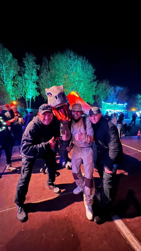 Drei Personen posieren fröhlich auf einem Festival, im Hintergrund festliche Lichter.