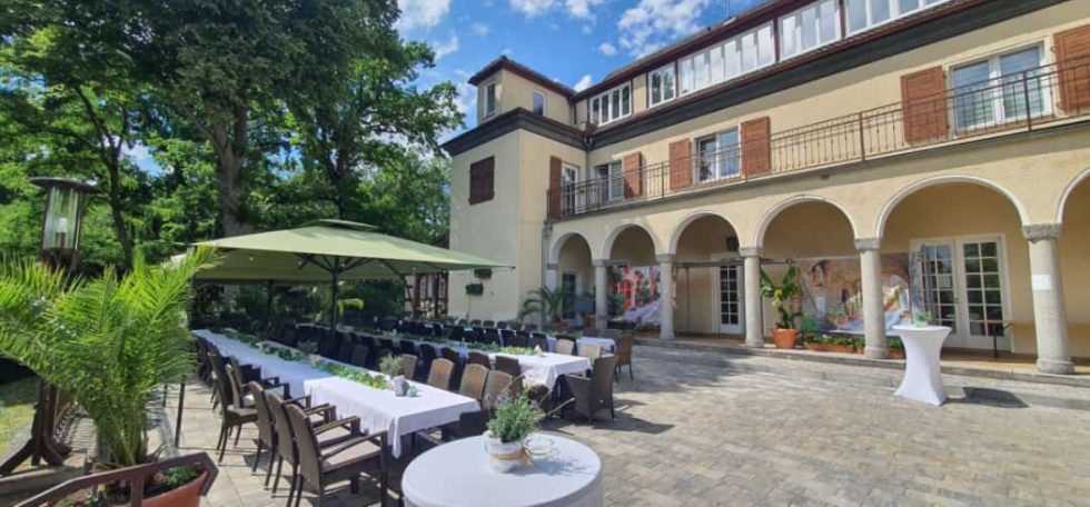 Helle Terrasse mit Tischen und Stühlen vor einem eleganten Gebäude im Freien.