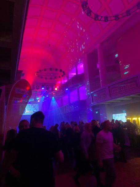 Bunte Lichter und Rauch im Raum mit zahlreichen Menschen während einer Feier.