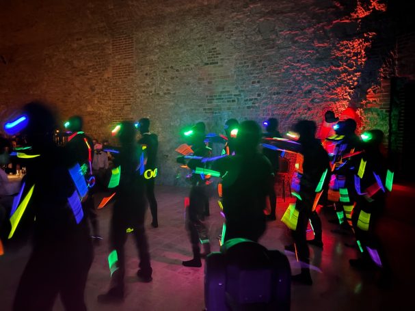 Menschen in schwarz gekleideten Anzügen mit leuchtenden Neonakzenten tanzen in einer bunten Umgebung.