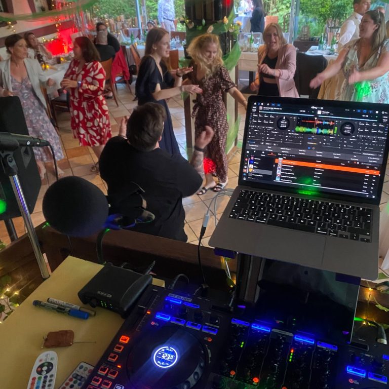 DJ-Pult mit Laptop, Tänzer in festlicher Atmosphäre im Hintergrund.
