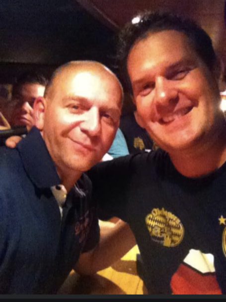 Zwei Männer lächeln lächelnd in einer Bar, einer trägt ein T-Shirt mit Aufdruck.